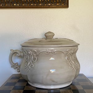 May include: A light beige ceramic chamber pot with a matching lid and a single side handle. The vessel features embossed scrollwork patterns around the upper rim and a smooth, glossy finish throughout the entire exterior surface.