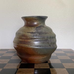 May include: A round ceramic vase with a wide, flared rim and a textured, bulbous body. The surface features a mottled glaze with blended tones of earthy brown, deep charcoal, and muted olive green, showcasing horizontal ridges along the sides.