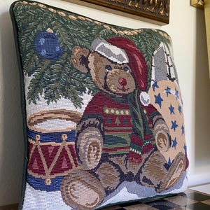 May include: A square tapestry throw pillow featuring a woven design of a brown teddy bear wearing a red Santa hat and a patterned sweater. The bear sits near a drum and a Christmas tree with blue ornaments against a background of stars.
