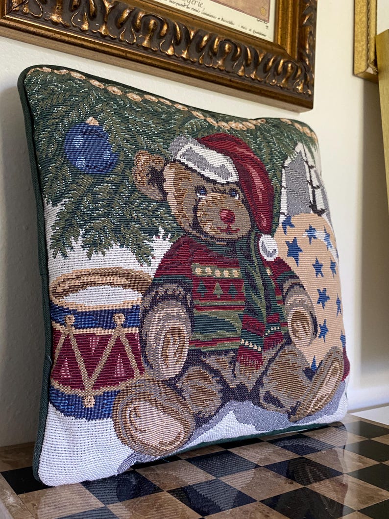 May include: A square tapestry throw pillow featuring a woven design of a brown teddy bear wearing a red Santa hat and a patterned sweater. The bear sits near a drum and a Christmas tree with blue ornaments against a background of stars.