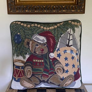 May include: A square tapestry throw pillow featuring a woven design of a brown teddy bear wearing a red Santa hat and a patterned sweater. The bear sits in front of a green pine tree with a blue ornament, a red drum, and a beige sack with blue stars.