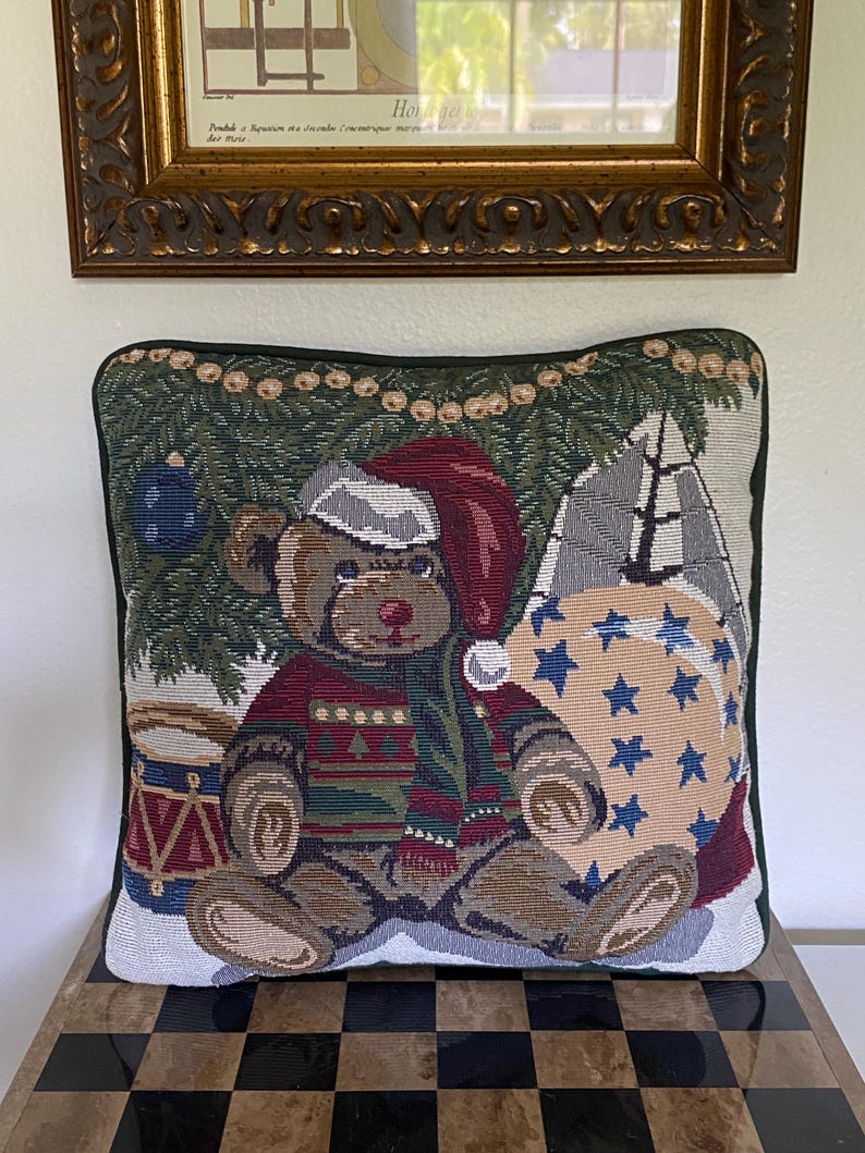 May include: A square tapestry throw pillow featuring a woven design of a brown teddy bear wearing a red Santa hat and a patterned sweater. The bear sits in front of a green pine tree with a blue ornament, a red drum, and a beige sack with blue stars.