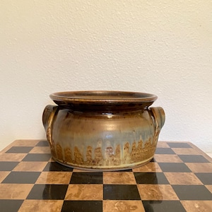May include: A round ceramic bowl with two side handles, featuring a glossy brown glaze at the top that transitions into a textured, dripped tan and beige finish toward the base. The vessel has a wide, open rim and a sturdy, low-profile design.