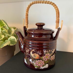 May include: A dark brown ceramic teapot with a glossy finish, featuring a curved spout, a matching lid, and a woven rattan swing handle. The body is decorated with a colorful painted scene of a peacock and pink peony flowers on a white rock.