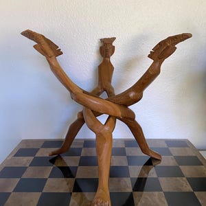 May include: A brown wooden sculpture featuring three interconnected figures with stylized heads and elongated limbs, arranged in a tripod formation. The figures are carved from a single piece of wood, with their legs intertwined to form a stable base.