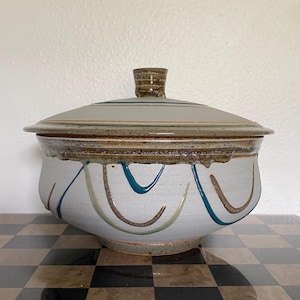 May include: A hand-thrown ceramic lidded bowl with a light gray matte finish. The bowl features hand-painted teal and brown curved line accents around the body, with a brown speckled glaze rim and a matching lid with a small knob handle.