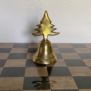 May include: A polished gold-colored metal hand bell featuring a stylized evergreen tree as the handle. The bell has a smooth, reflective surface and a flared base, with the tree silhouette extending upward from the top of the bell structure.