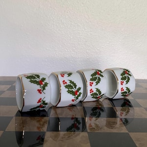May include: A set of four white ceramic napkin rings, each featuring a painted pattern of green holly leaves and bright red berries. The edges of each cylindrical napkin ring are finished with a thin, metallic gold trim.