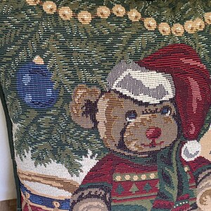 May include: A square throw pillow featuring a woven tapestry design of a teddy bear wearing a red Santa hat and a patterned sweater. The bear sits near a decorated Christmas tree with a blue ornament and a string of lights against a dark green background.