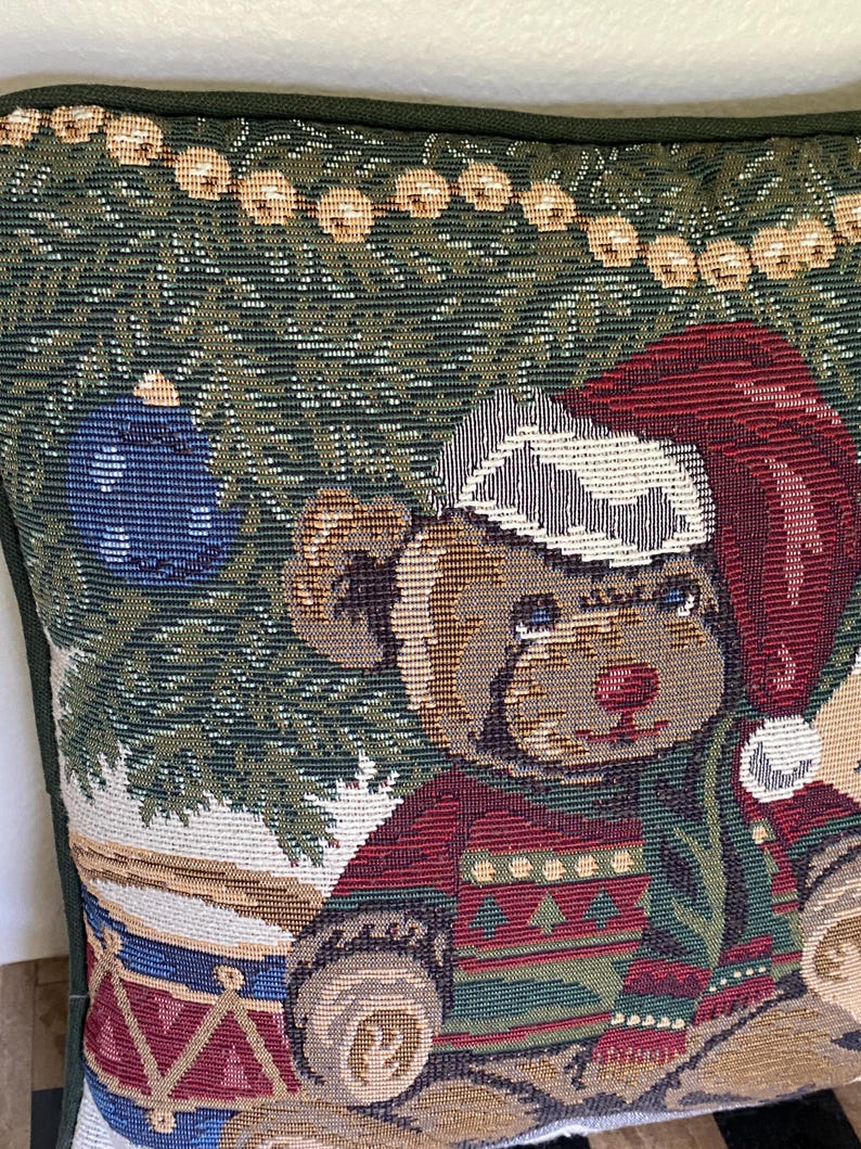 May include: A square throw pillow featuring a woven tapestry design of a teddy bear wearing a red Santa hat and a patterned sweater. The bear sits near a decorated Christmas tree with a blue ornament and a string of lights against a dark green background.