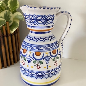 May include: A white ceramic pitcher with blue, orange, and yellow hand-painted designs. The pitcher features a handle and spout, with a decorative pattern around the top and base. The design includes floral and geometric elements, creating a vibrant and artistic look.