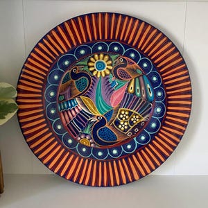 May include: A round, hand-painted decorative plate featuring a vibrant bird and floral design. The plate has a dark blue center with colorful birds, flowers, and leaves. The outer edge is orange with vertical stripes, creating a striking contrast.