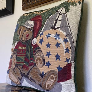 May include: A square decorative throw pillow featuring a woven tapestry design of a brown teddy bear wearing a red Santa hat and a green patterned sweater. The bear sits next to a large beige circular ornament decorated with blue stars.