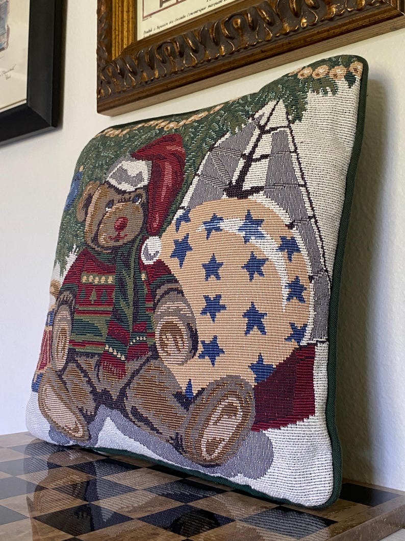 May include: A square decorative throw pillow featuring a woven tapestry design of a brown teddy bear wearing a red Santa hat and a green patterned sweater. The bear sits next to a large beige circular ornament decorated with blue stars.