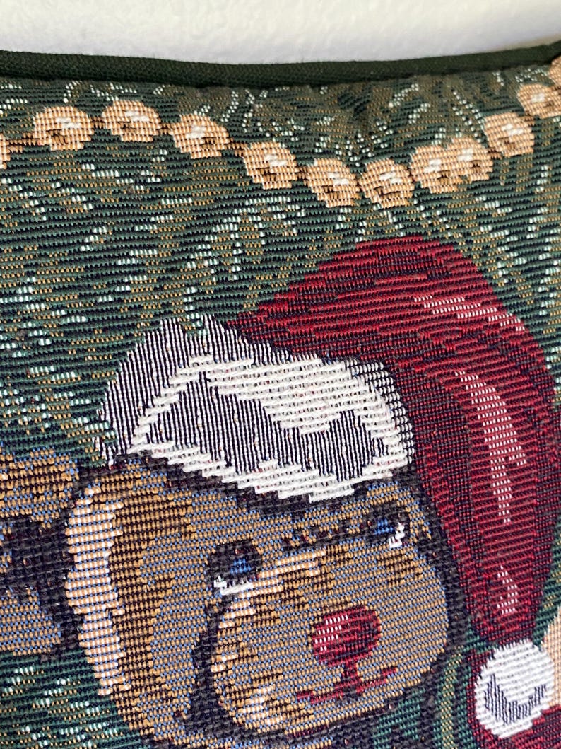 May include: A woven tapestry pillow cover featuring a brown bear wearing a red Santa hat with a white pom-pom and trim. The background is a dark green fabric with a pattern of light-colored circular ornaments arranged in a horizontal row.