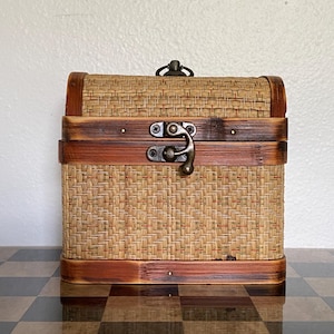 May include: A rectangular storage chest with a domed lid, featuring woven tan wicker panels framed by dark brown stained wood trim. The chest includes a central antique brass-colored metal latch and a small matching handle on the top lid.