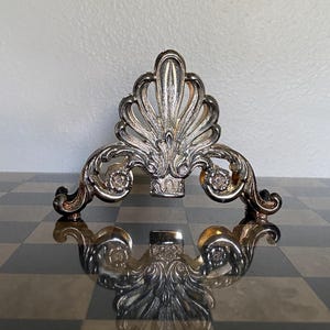 May include: A silver-toned metal napkin holder featuring an ornate, symmetrical design with a central fan-like motif and curved scrollwork accents on both sides. The piece has a polished, reflective finish and a wide, stable base structure.