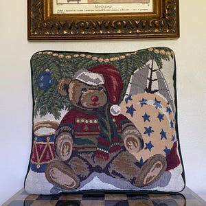 May include: A square tapestry throw pillow featuring a woven design of a brown teddy bear wearing a red Santa hat and a patterned sweater. The bear sits before a green pine tree, a drum, and a large beige sack decorated with blue stars.