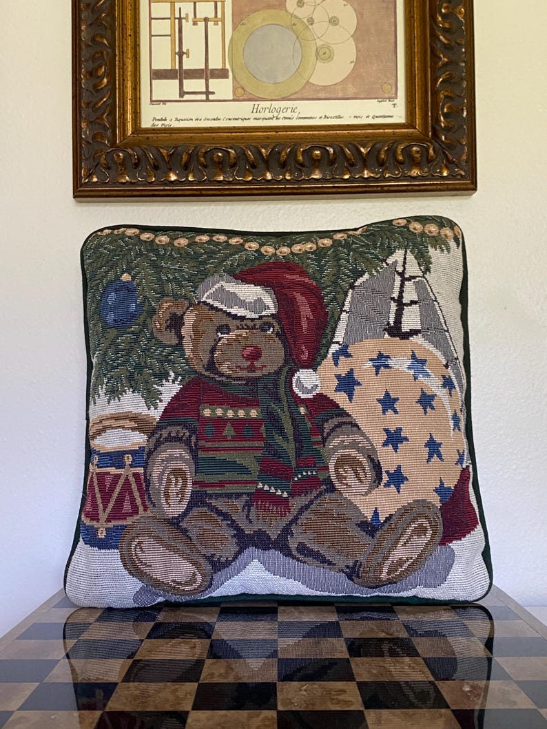 May include: A square tapestry throw pillow featuring a woven design of a brown teddy bear wearing a red Santa hat and a patterned sweater. The bear sits before a green pine tree, a drum, and a large beige sack decorated with blue stars.