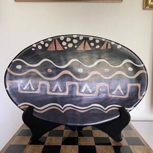 May include: An oval-shaped black ceramic platter featuring hand-painted geometric patterns in tan and white. The design includes horizontal wavy lines, triangles, and circular dots. The platter is displayed on a black plastic stand.