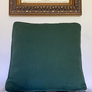 May include: A square throw pillow in a solid dark forest green color. The pillow features a simple, clean design with piped edges along the perimeter, providing a structured look. The fabric appears to have a matte, woven texture throughout.