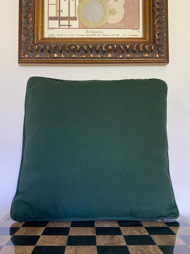 May include: A square throw pillow in a solid dark forest green color. The pillow features a simple, clean design with piped edges along the perimeter, providing a structured look. The fabric appears to have a matte, woven texture throughout.