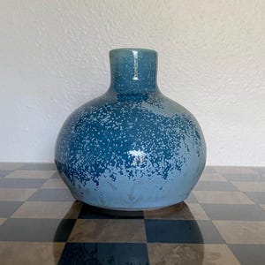 May include: A small, round ceramic vase with a narrow neck and a glossy finish. The surface features a gradient of deep blue at the top transitioning to a lighter, pale blue at the base, with a speckled, mottled texture throughout the glaze.