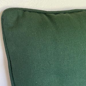 May include: A square throw pillow cover in a solid dark forest green color. The fabric features a visible woven canvas texture with a piped edge detail along the seam, creating a structured border around the perimeter of the cushion cover.