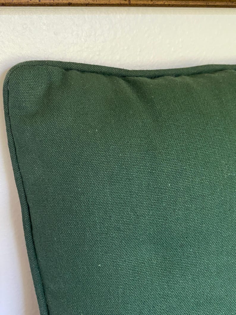 May include: A square throw pillow cover in a solid dark forest green color. The fabric features a visible woven canvas texture with a piped edge detail along the seam, creating a structured border around the perimeter of the cushion cover.