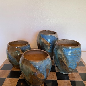 May include: Four rounded ceramic cups with a speckled brown interior and a reactive glaze exterior featuring swirling patterns of deep blue, tan, and dark brown. Each cup has a wide, open mouth and a tapered base, showcasing a unique, hand-thrown appearance.