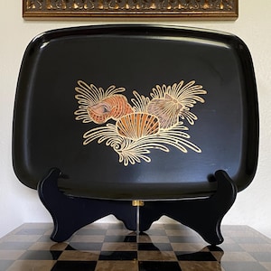 May include: A rectangular black tray with rounded corners featuring a central gold and orange illustration of three seashells surrounded by stylized golden seaweed or coral patterns. The tray is displayed on a black stand for viewing.