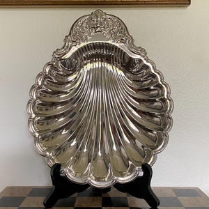 May include: A polished silver-tone metal serving dish shaped like a large scallop shell. The dish features deep, radiating fluted grooves and an ornate, embossed decorative crest at the top edge, supported by a black plastic display stand.