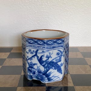 May include: A hexagonal ceramic planter featuring a blue and white underglaze design of a bird perched on a flowering branch. The rim is finished with a thin brown band, and the exterior panels display intricate geometric and floral patterns.