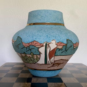 May include: A ceramic vase with a speckled light blue finish, featuring a desert landscape scene with cacti, rock formations, and two figures in white robes. The design includes horizontal gold bands near the top and bottom rims.