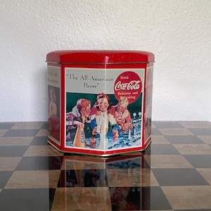 May include: A hexagonal red metal tin featuring a vintage Coca-Cola advertisement. The front panel displays the text "The All-American Pause" and "Drink Coca-Cola Delicious and Refreshing" above an illustration of three people at a soda fountain.