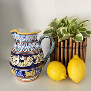 May include: A decorative ceramic pitcher with a blue, yellow, and orange floral design. The pitcher has a curved handle and a spout. Two yellow lemons and a potted plant in a bamboo holder are next to the pitcher.