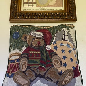 May include: A square decorative throw pillow featuring a woven tapestry design of a brown teddy bear wearing a red Santa hat and a patterned sweater. The bear sits near a toy drum and a large sack decorated with blue stars against a pine tree background.