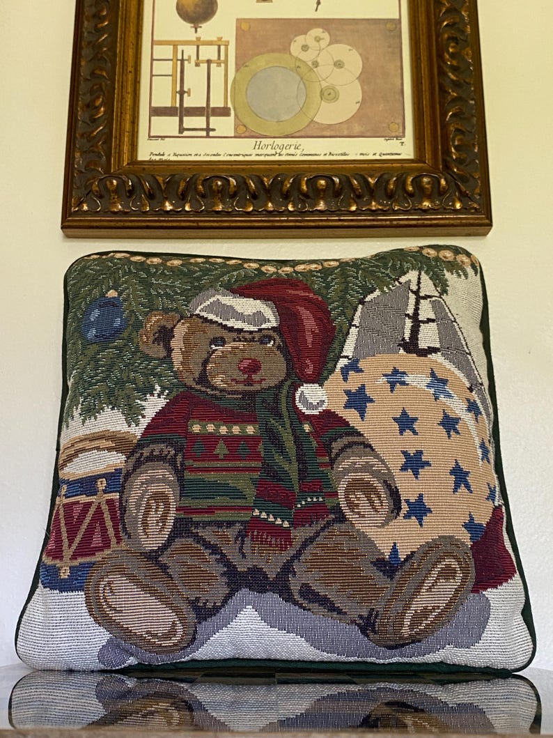 May include: A square decorative throw pillow featuring a woven tapestry design of a brown teddy bear wearing a red Santa hat and a patterned sweater. The bear sits near a toy drum and a large sack decorated with blue stars against a pine tree background.