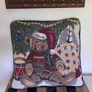 May include: A square decorative throw pillow featuring a woven tapestry design of a brown teddy bear wearing a red Santa hat and a patterned sweater. The bear sits near a Christmas tree, a blue drum, and a large beige sack decorated with blue stars.