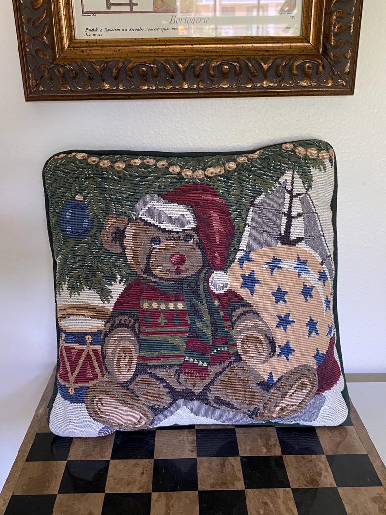 May include: A square decorative throw pillow featuring a woven tapestry design of a brown teddy bear wearing a red Santa hat and a patterned sweater. The bear sits near a Christmas tree, a blue drum, and a large beige sack decorated with blue stars.