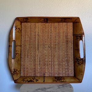 May include: A square serving tray constructed from natural bamboo with a woven base. The tray features a glossy finish with dark, mottled brown markings on the frame and integrated side handles for carrying. The center has a tight, light brown woven texture.