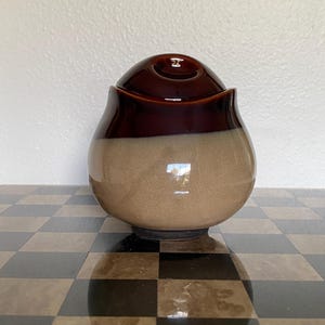 May include: A ceramic jar with a rounded body and a matching lid. The upper portion of the jar and the lid are finished in a glossy dark brown glaze, while the lower portion features a smooth, matte-textured light beige finish.