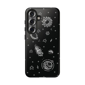 May include: Black phone case featuring a white space-themed design. The case includes illustrations of a sun, stars, planets, a rocket, and a UFO. The phone case has a camera module on the upper left side.