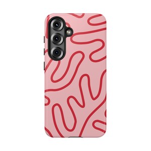 May include: A pink phone case with a red abstract squiggly line design. The case has a camera module with three lenses and a flash. The phone case is made of a hard plastic material.