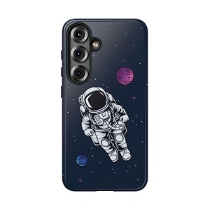 May include: A navy blue phone case featuring a detailed illustration of an astronaut floating in space. The design includes planets, stars, and a dark blue background. The case has a camera module on the top left.