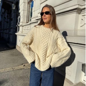 May include: A cream-coloured cable-knit jumper with a round neckline. The jumper features a textured pattern and long sleeves. The person is wearing blue jeans and sunglasses. The jumper is a cosy and stylish garment.