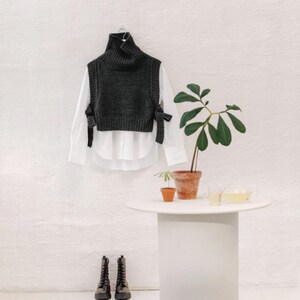 May include: A dark gray turtleneck sweater vest with bow details, layered over a white button-down shirt. Black combat boots are on the floor. A small white table holds a potted plant and a glass of liquid.