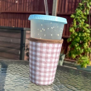 May include: A clear plastic tumbler with a light blue lid and a transparent straw, featuring small white flecks throughout the cup. The tumbler is encased in a fabric sleeve with a light pink and white gingham check pattern and brown trim.