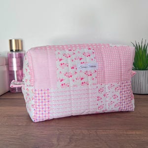 Peut inclure: Une pochette matelassée rectangulaire présentant un motif patchwork de tissus roses et blancs. Les motifs incluent des carreaux vichy, des fines rayures et de petites fleurs. Une étiquette en tissu blanc avec le texte Serene Stitches est cousue.