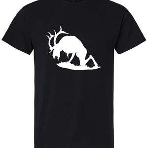 May include: Black t-shirt featuring a white silhouette of a deer with large antlers, bent over as if grazing. The deer is positioned on a patch of grass. The shirt is made of a soft material.
